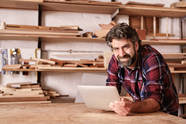 Portrait of a small business owner smiling at the camera, resting on his workbench and holding a digital tablet, with shelves of wood behind him in his woodwork studio